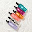 Top-down view of six translucent, colorful Pion After Shave Cologne bottles lined up diagonally. The colors include teal, magenta, orange, coral, clear, and purple. The bottles rest on a bright white background covered by a thin layer of rippling water, creating clean reflections and shadows.